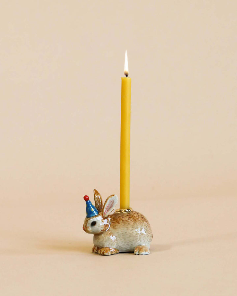 A hand-painted Rabbit/Bunny Cake Topper wearing a small blue party hat, supporting a lit yellow candle on its back, set against a plain beige background.