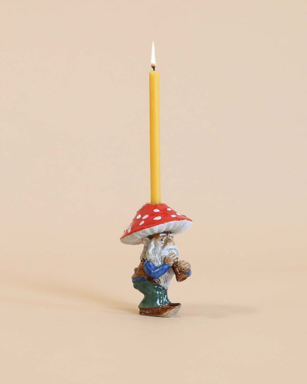 A whimsical Mushroom Gnome Cake Topper, hand-painted and crafted from fine porcelain, with a red and white mushroom cap, holding a lit yellow candle, set against a plain beige background.