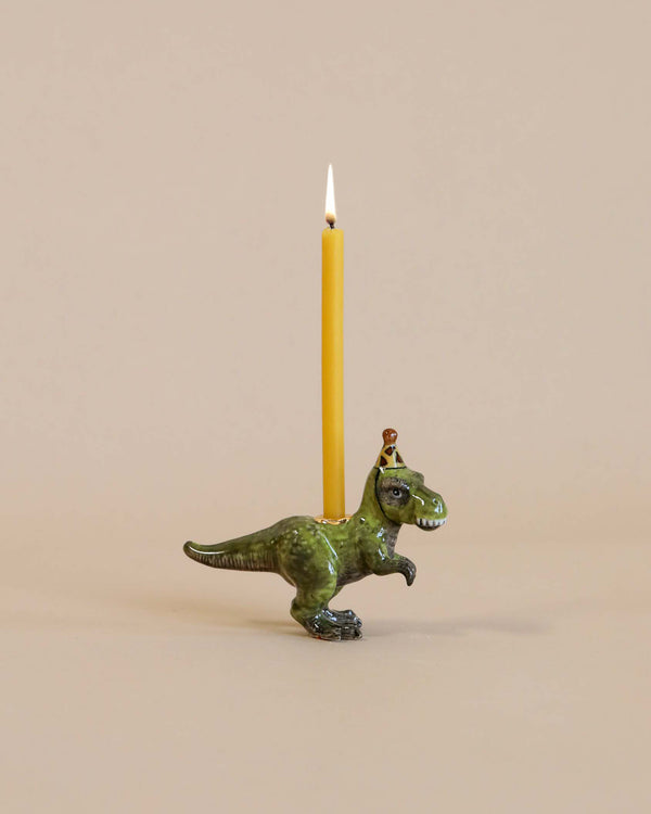 A whimsical T-Rex "Party Animal" Cake Topper shaped like a green t-rex dinosaur wearing a tiny golden crown, holding a lit yellow candle on its back against a beige background. This hand-painted porcelain keepsake adds