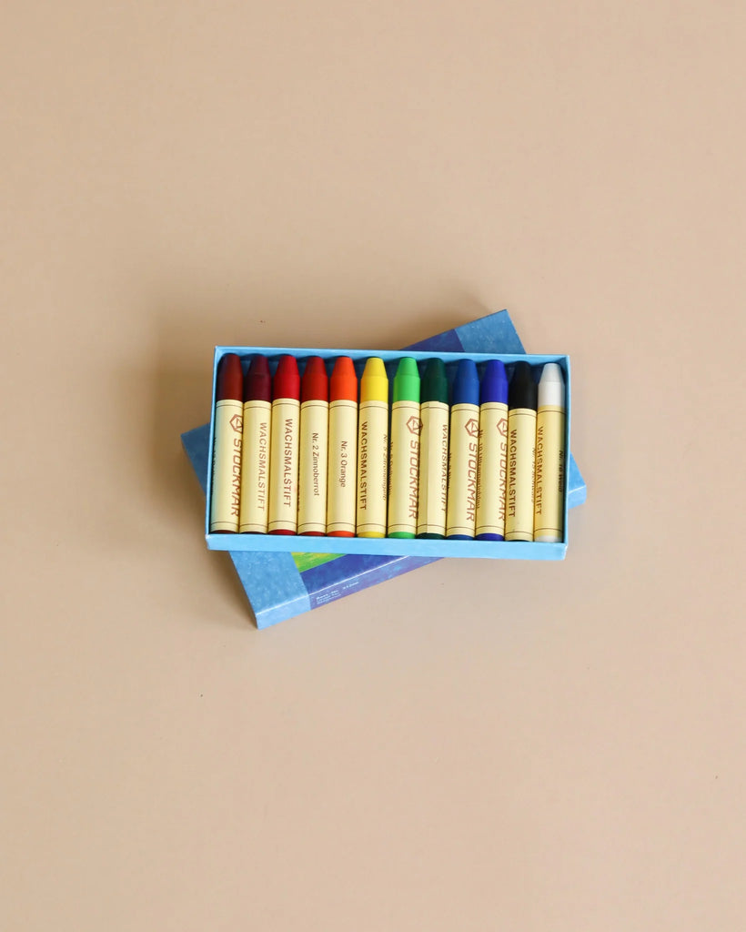 The Stockmar Crayon Set features a non-toxic, vibrant 10-color collection, including yellow, orange, red, purple, green, blue, and black. The crayons are neatly arranged in a wooden organizer tray on a brown surface to meet any artistic demands.