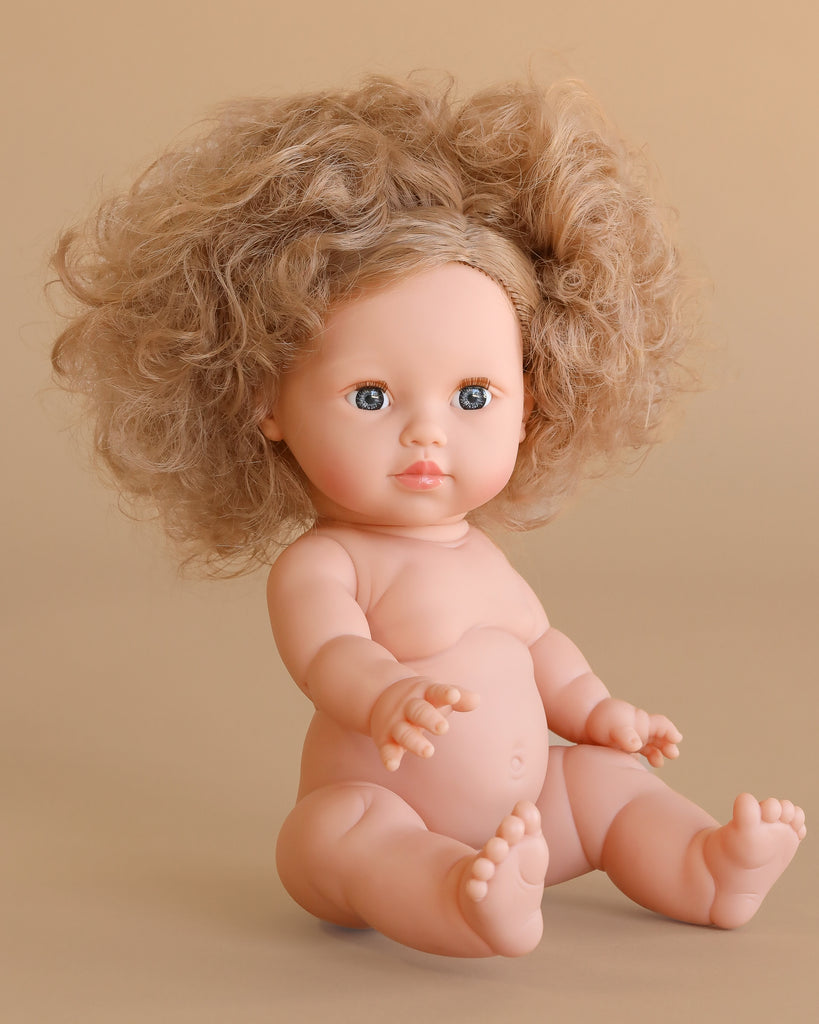 The Minikane Baby Doll (13") - Lola sits on a beige background with curly light brown hair, blue eyes, and a hard body. Lola features realistic details and a charming expression. Clothing sold separately.