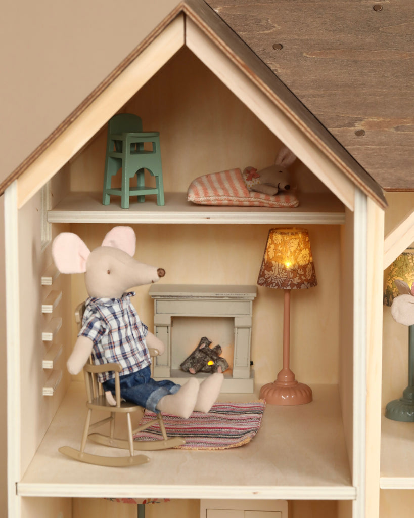 A cozy Maileg Farmhouse - Fully Furnished dollhouse scene featuring a Maileg collection doll mouse in a checkered shirt, sitting in a rocking chair by a fireplace, relaxing with a book. A small lamp, a cushion