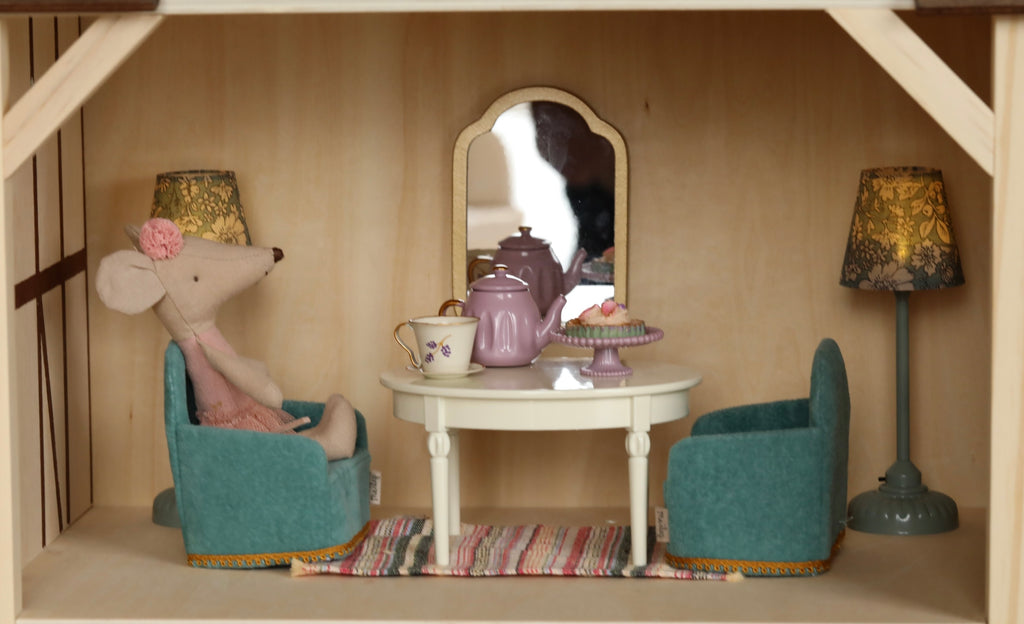 A charming scene from the Maileg Farmhouse - Fully Furnished collection depicting a toy mouse sitting at a small table with teapot and cupcake, inside a wooden dollhouse, with a mirror and two lamps.