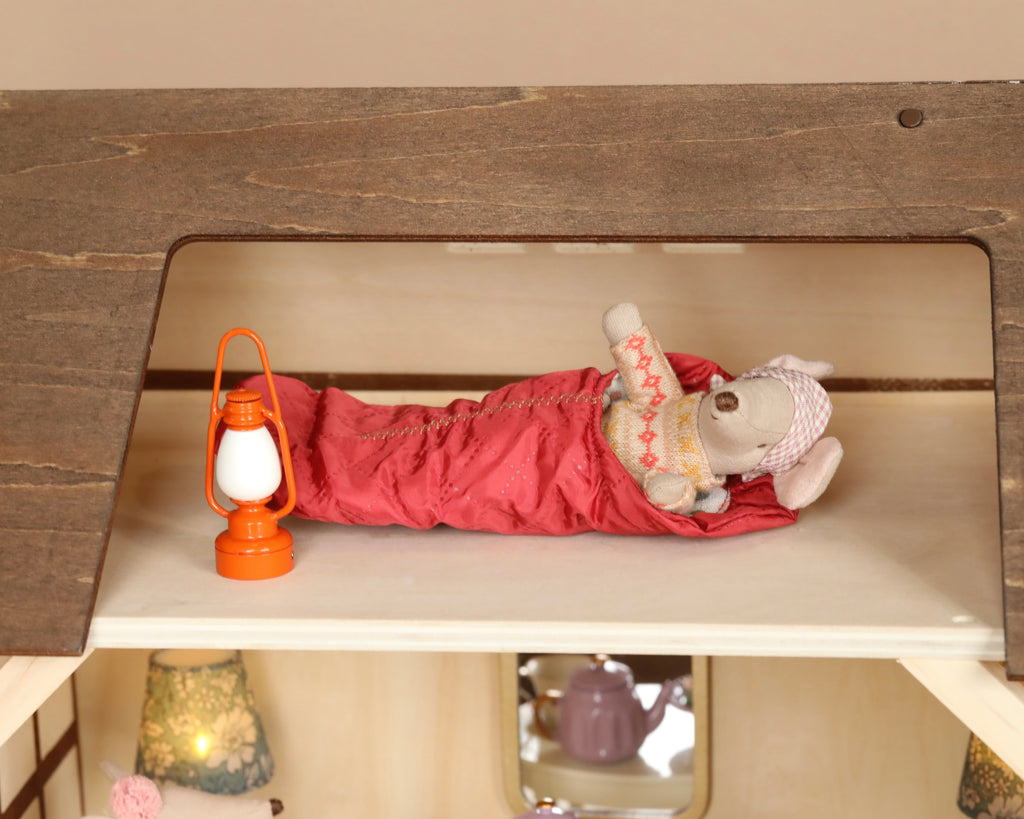 A Maileg Farmhouse - Fully Furnished with a plush teddy bear wearing a nightcap and sleeping bag lies on a wooden shelf with an orange lantern beside it, evoking the cozy charm of a farmhouse.
