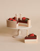 A wooden cake stand displaying a Handmade Chocolate Layer Cake On A Stand divided into slices, each topped with a red apple and white candles, on a beige background. The toy is crafted from non-toxic paint.