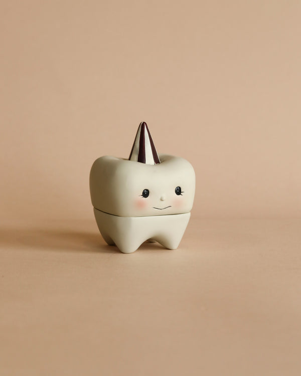 A small Konges Sløjd Stone Tooth Box shaped like a tooth, with a cute, smiling face and painted blush cheeks. Its top resembles a dark and white striped party hat. Designed to hold a child's first teeth, the container sits against a simple, solid beige background.
