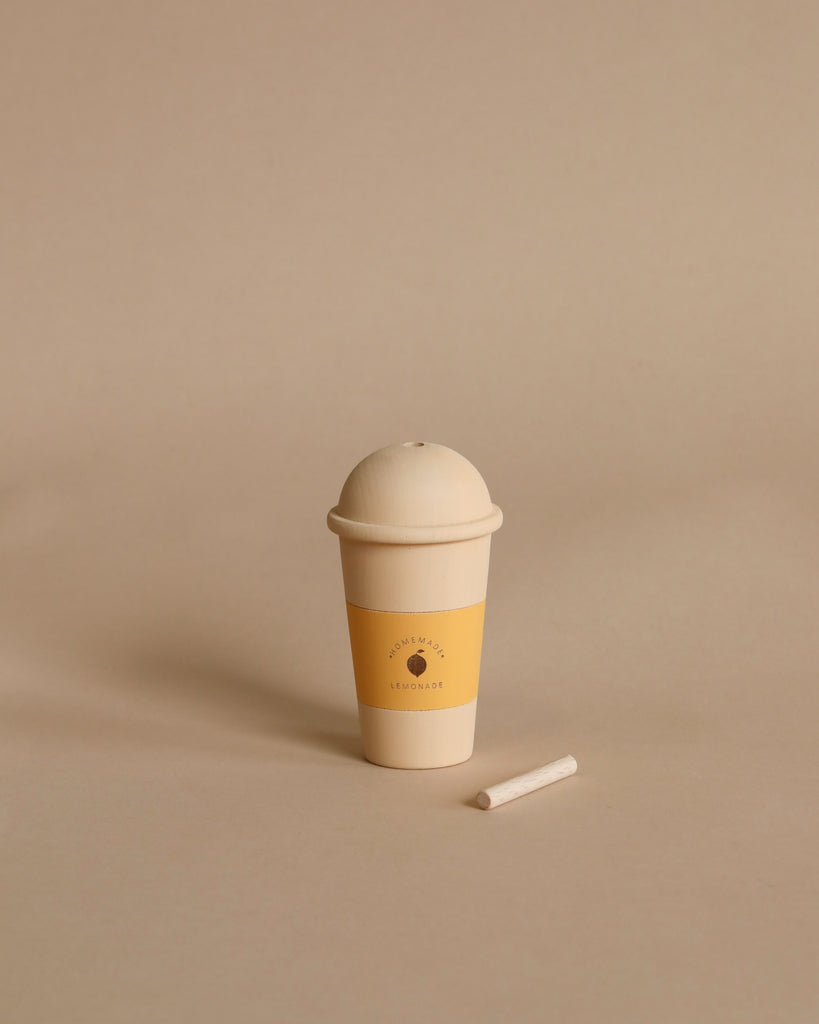 A minimalist image of a Handmade Wooden Lemonade with a cream-colored dome lid next to a small white piece of chalk, all set against a plain beige background. The cup and chalk feature non-toxic water-based paint.