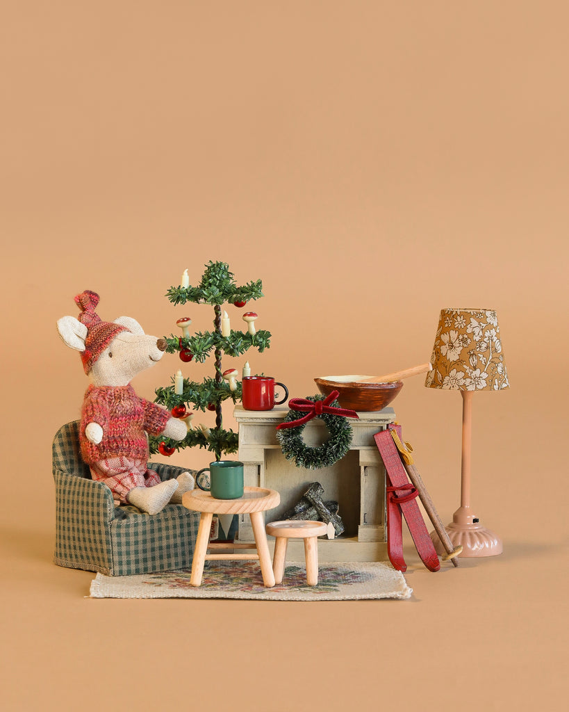 The Maileg Cozy Christmas Set features a winter ski mouse in a red sweater by a mini fireplace, with a tiny Christmas tree, coffee table, mugs, book, and a vintage floor lamp, all on a warm beige background.