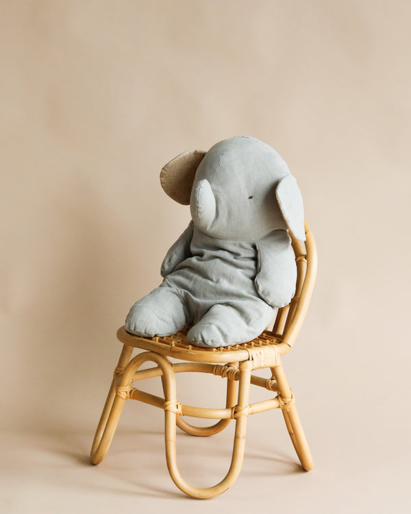 A Maileg Big Grey Elephant soft toy sitting on a small rattan chair against a plain beige background. The toy is facing away, showing its back and large floppy ears.