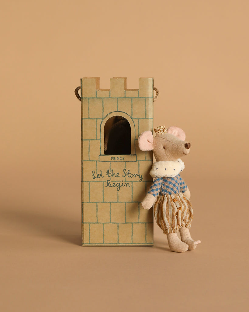 The Maileg Prince Mouse, Big Brother—a small cotton mouse in royal clothes and a crown—leans on a cardboard castle tower with a window and "Let the Story Begin" written on it. The background is light brown.
