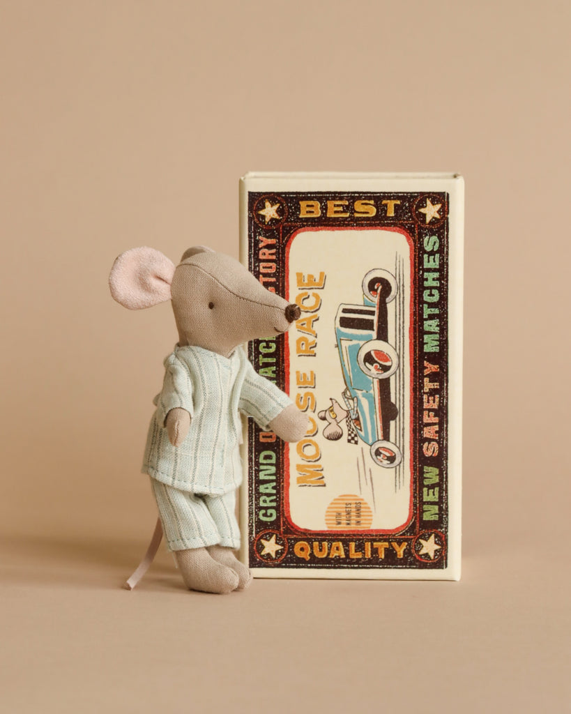 A small Maileg Big Brother mouse dressed in a pale blue outfit stands next to an oversized vintage matchbox bed with colorful graphics, inspecting the design. The background is a neutral beige.