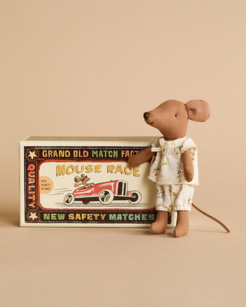 A small Maileg Big Sister, Mouse In Matchbox in white pajamas stands beside a vintage matchbox bed that reads "Mouse Race" with an image of a red race car. The soft fabrics of the plush mouse contrast the beige background while the matchbox text includes "Grand Old Match Factory" and "New Safety Matches".