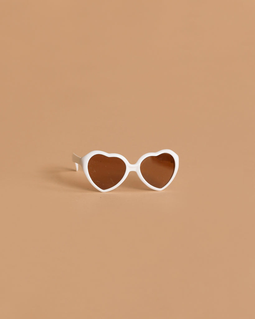 Minikane Doll Accessories | Loona Cream Heart Sunglasses: cream heart-shaped doll glasses with dark lenses, ideal for Minikane dolls, displayed on a solid light brown background.