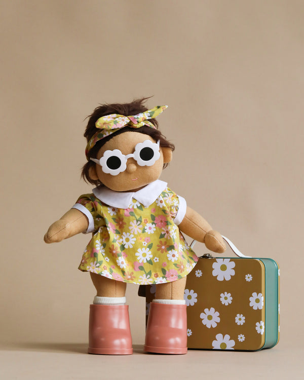 A plush Dinkum Doll - Sprout (Extended Pack) dressed in a floral dress, headband, and white-rimmed sunglasses stands beside a matching floral suitcase. The doll also wears pink boots, showcasing a charming unisex outfit. The background is a neutral beige color.