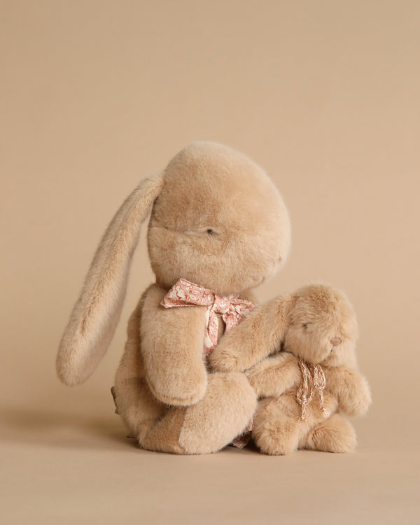 Two Maileg Plush Bunny Mom & Baby toys, one large with a pink bow and one small, sitting together against a tan background, with the larger one seeming to comfort the smaller one.