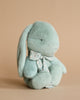 A Maileg Small Plush Bunny - Mint with long ears and a floral bow around its neck, sitting against a plain beige background. Its eyes appear closed and its overall color is soft blue.