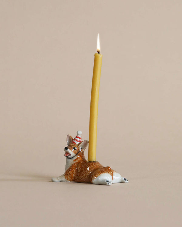 A Corgi Cake Topper shaped like a reclining fox, crafted from hand-painted porcelain, supports a tall, lit yellow candle against a plain beige background.