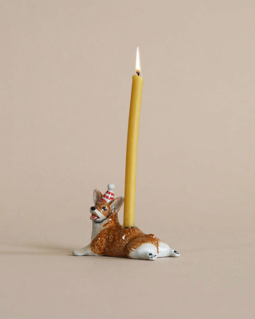 A Corgi Cake Topper shaped like a reclining fox, crafted from hand-painted porcelain, supports a tall, lit yellow candle against a plain beige background.