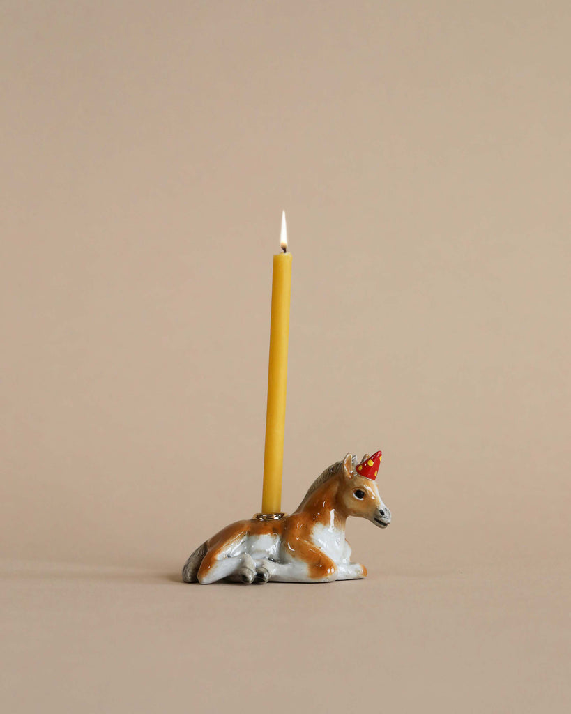 A lit yellow candle is inserted into a hand-painted porcelain holder shaped like a horse with a painted red bow on a beige background.