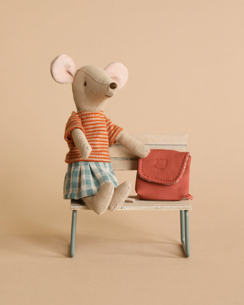 A plush mouse with a light brown body sits on a small white bench. The Maileg Big Sister With Backpack - Old Rose is wearing a red-striped shirt and blue checkered shorts. Next to her is a small red bag, making her the perfect kindergarten toy. The background is a plain beige color.