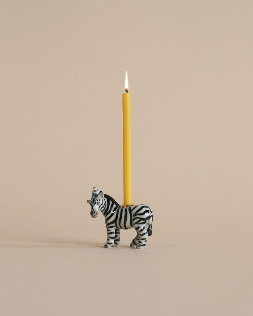 A small hand-painted Zebra Cake Topper with a lit yellow candle inserted on its back, standing against a plain, light beige background.