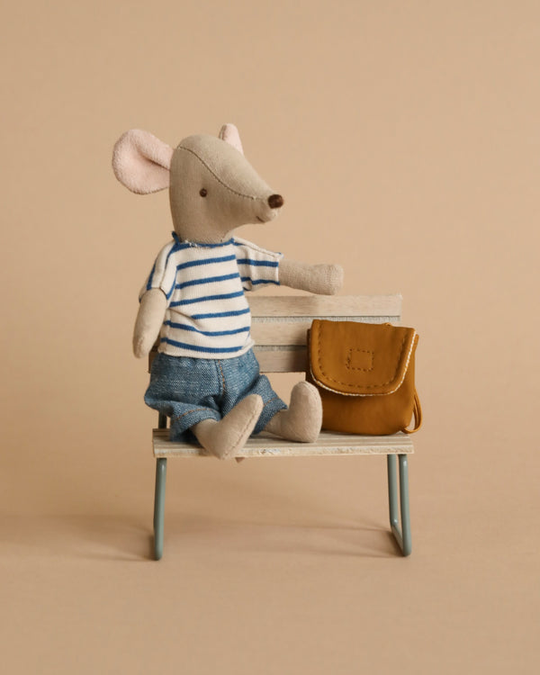 A Maileg Big Brother With Backpack toy dressed in a striped shirt and denim shorts, sitting on a small wooden bench with a stack of books and a tiny brown bag, against a beige background.