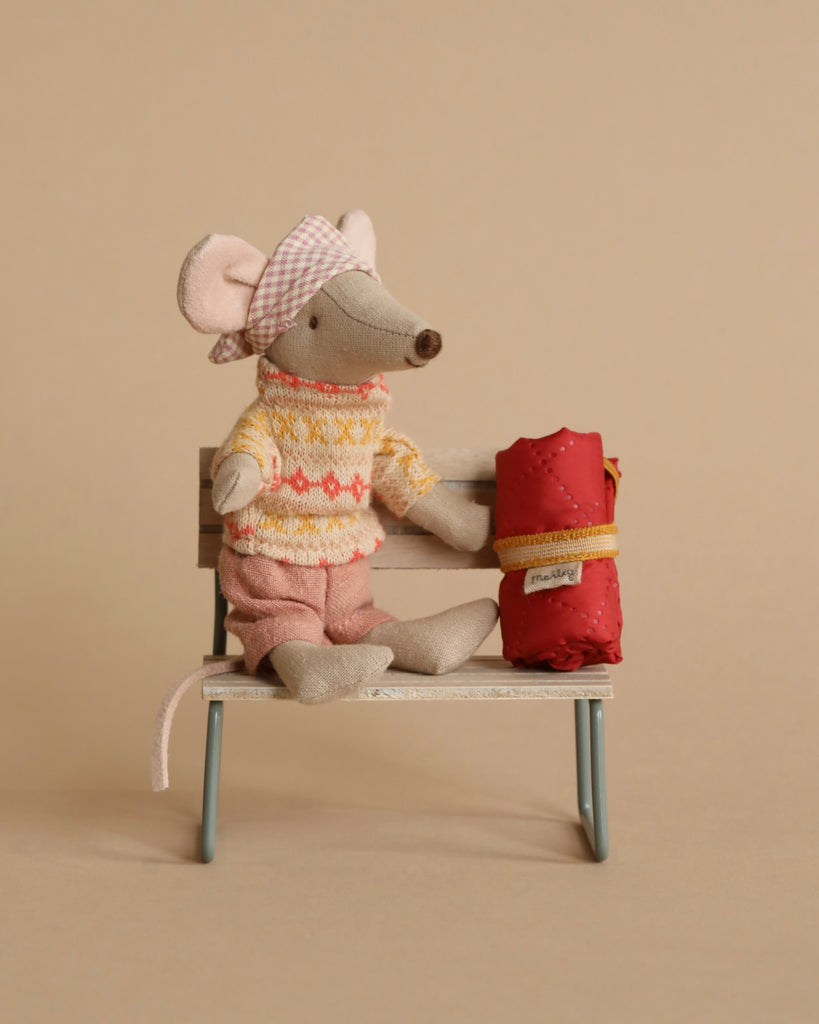 A small, stuffed mouse toy wearing a knitted sweater, pink shorts, and a pink gingham hat sits on a miniature bench. The Maileg Hiker Mouse, Big Sister is complemented by a red rolled-up sleeping bag secured with a yellow strap. The background is a plain beige color.