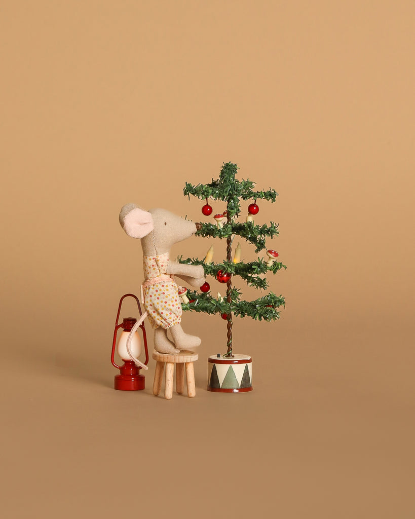 A Maileg mouse sits on a Maileg table and stool, decorating a miniature Christmas tree with red ornaments from the Maileg Holiday Tree Decorating Set. A tiny red lantern completes the festive scene against a beige background.