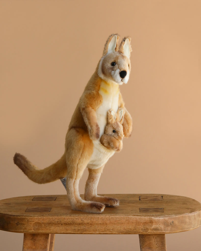 The Kangaroo Mom Stuffed Animal, featuring a baby joey in its pouch and crafted in the style of HANSA animals, stands on a wooden stool against a plain beige background.