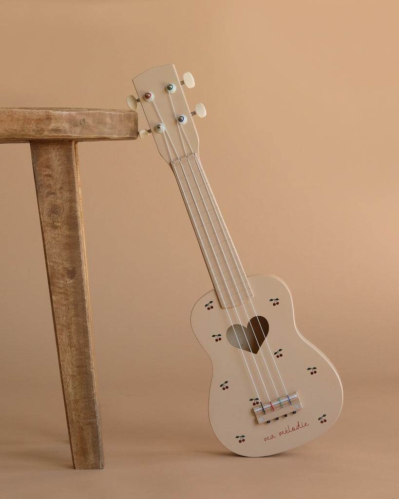 The Wooden Ukulele - Cherry, a charming cream-colored instrument with a heart-shaped sound hole and paw print accents, leans against a wooden stool on a beige background—an adorable, eco-friendly choice for music lovers.
