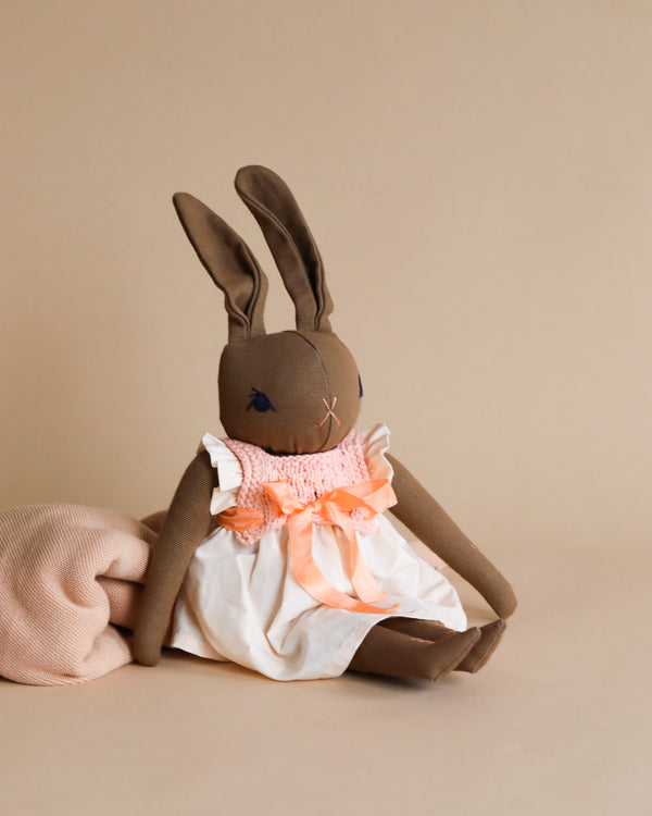 A handmade Polka Dot Club Large Brown Rabbit in Hand Knit Dress with long ears, wearing a white dress with a peach-colored bow, sits against a neutral beige background. The toy is made from cotton canvas and is hand embroidered.