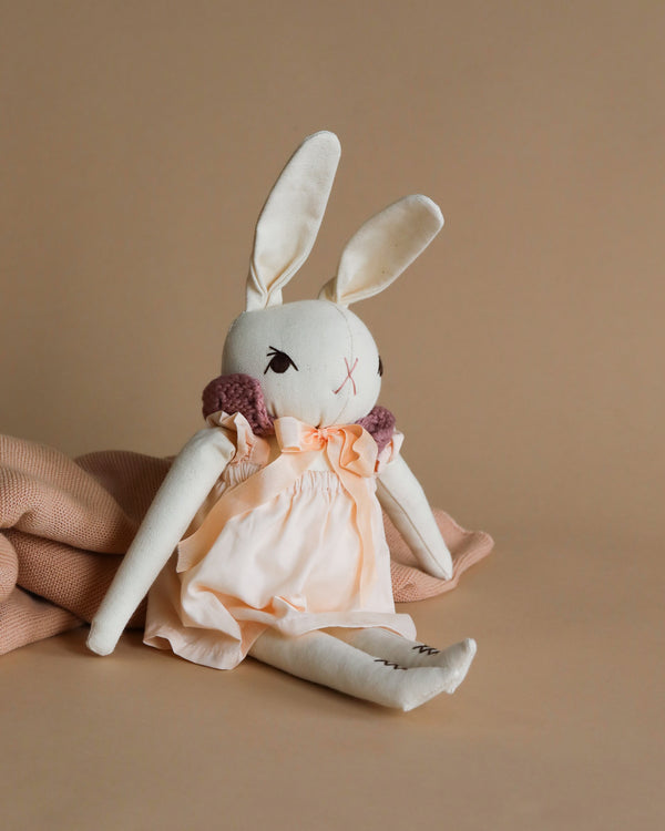 A Polka Dot Club Medium Cream Rabbit with Hand Knit Collar toy wearing a peach dress, seated against a beige backdrop with a textured cloth draped beside it.