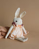 A Polka Dot Club Medium Cream Rabbit with Hand Knit Collar toy wearing a peach dress, seated against a beige backdrop with a textured cloth draped beside it.