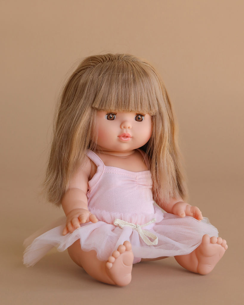 The Minikane Baby Doll (13") - Nina with Clothing features long, straight light brown hair and bangs. Made in Spain, this anatomically correct doll wears a pale pink dress with a tulle skirt and sits barefoot against a beige background.