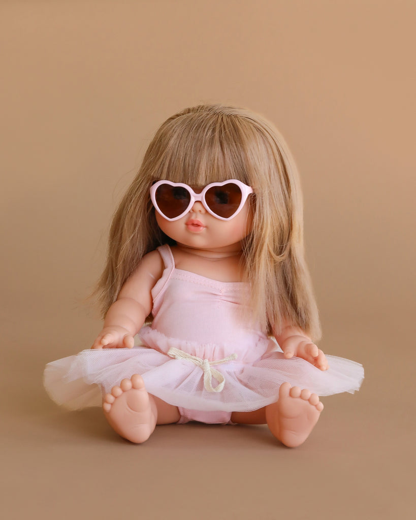The Minikane Baby Doll (13") - Nina with Clothing, featuring long light brown hair, heart-shaped sunglasses, and a pink ballerina tutu, is an adorable anatomically correct doll made in Spain, shown against a beige background.