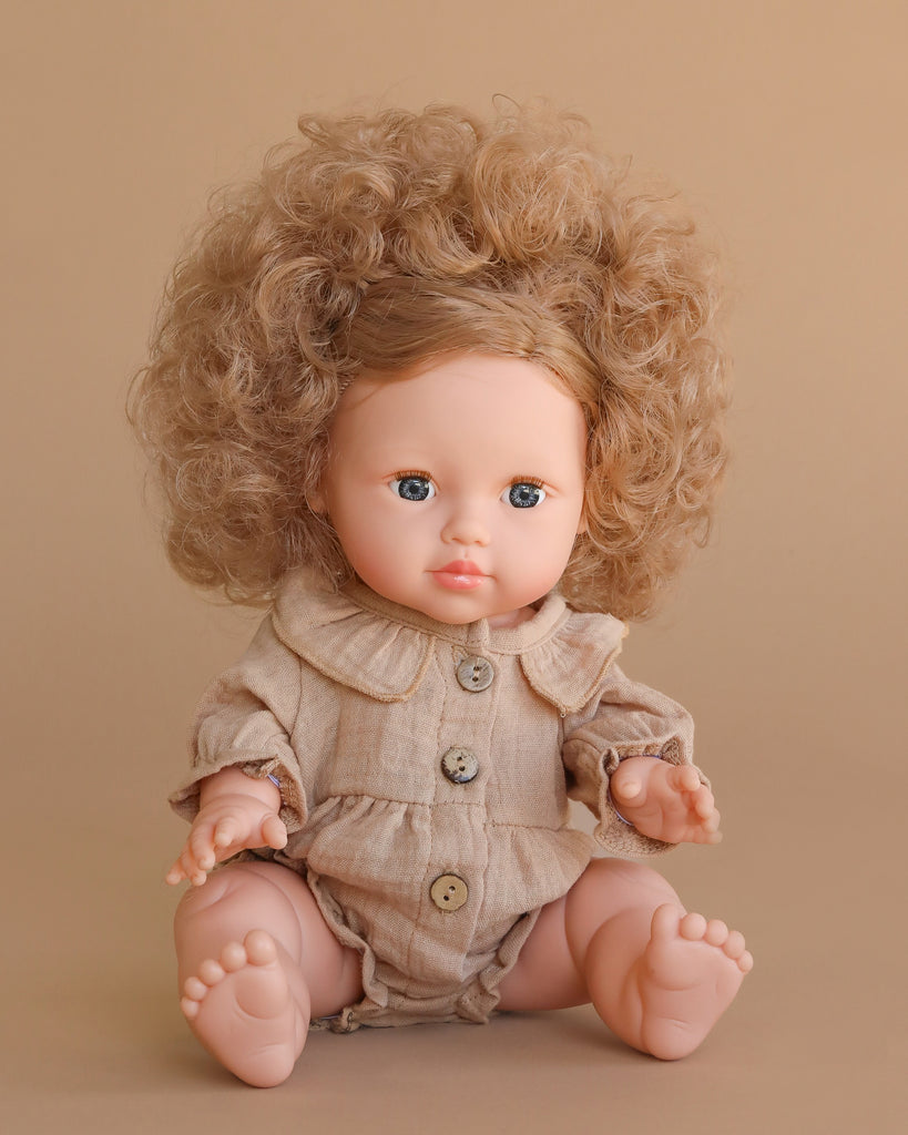 The Minikane Baby Doll (13") - Lola with Clothing features curly hair, blue eyes, and a beige outfit with large buttons. This anatomically correct doll is beautifully crafted in Spain and sits against a plain beige background.