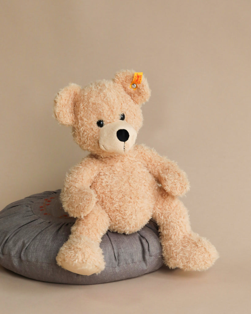 A beige Steiff Fynn Teddy bear sitting on a gray cushion against a light brown background, with the Button in Ear trademark visible on its ear.