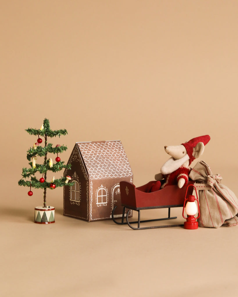 The Maileg Santa Mouse Christmas Set features Santa Mouse, a red sleigh, a mouse gingerbread house, a striped sack, and a red lantern, all arranged on a beige background.