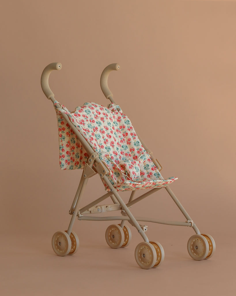 The Konges Sloejd Doll Stroller - Lumina, shown against a light brown background, features a beige frame, floral-patterned removable fabric seat, four sets of double wheels, and curved handles.