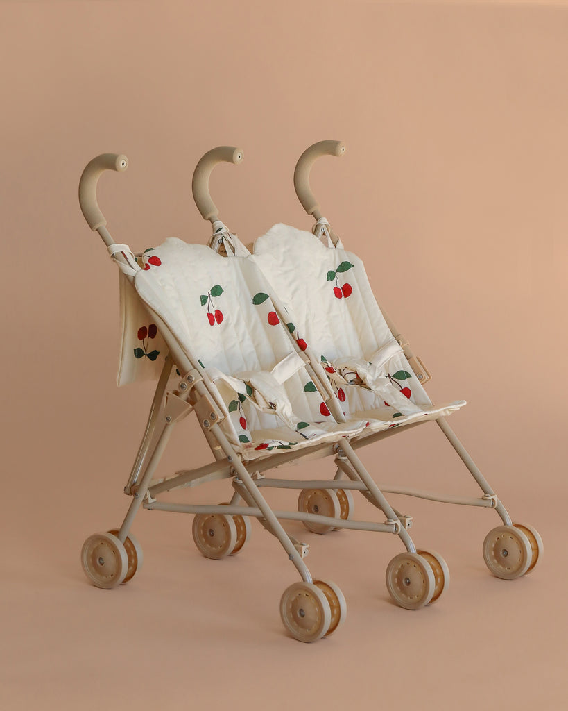 The Konges Sloejd Twin Doll Stroller - Cherry features a beige frame, light brown wheels and handles, two seats with white fabric adorned in red cherry patterns, and secure harnesses, shown against a plain beige background.
