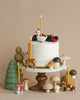 A whimsical cake decorated with forest-themed elements including mushrooms, a stag cake topper, a fox, and a hen, surrounded by green and brown fine porcelain trees and tall candles on a neutral backdrop.
