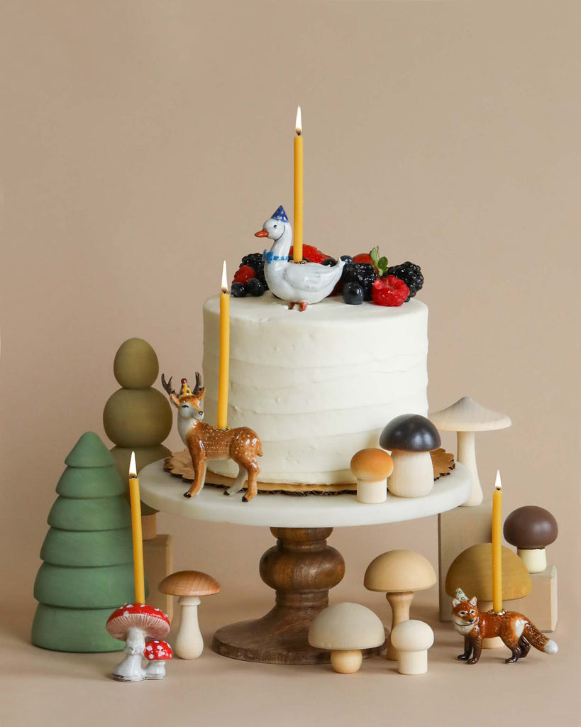 A whimsical, heirloom-quality birthday Mushroom Cake Topper on a wooden stand, adorned with berries, a fine porcelain hen on top, and surrounded by decorative mushrooms, trees, and animal figurines.