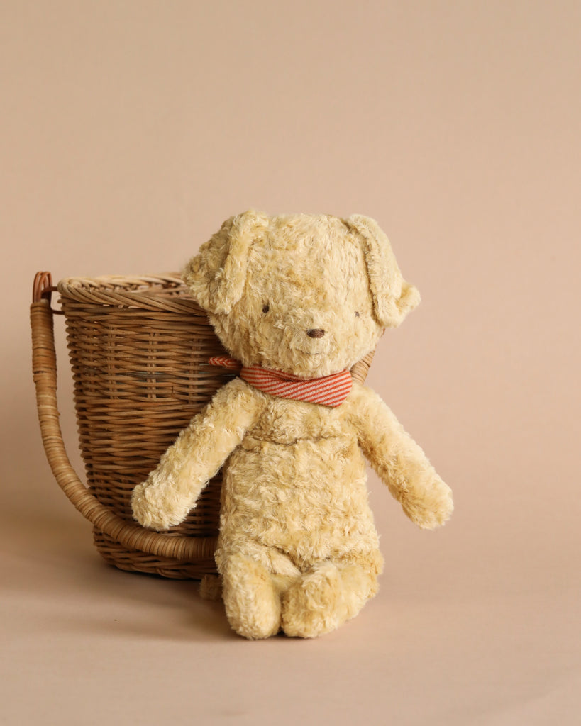 An old, worn Maileg Plush Dog with a striped bow tie sits in front of a wicker basket against a beige background, evoking a vintage dog aesthetic.