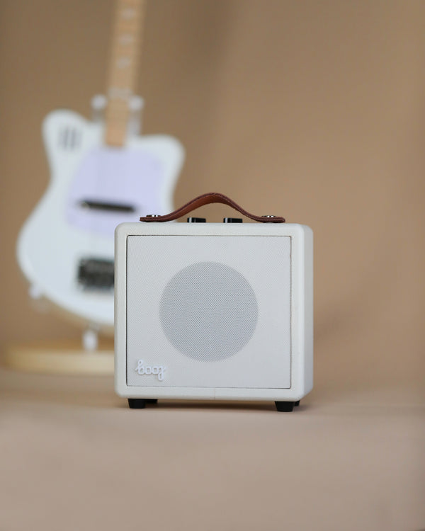 A small white Mini Guitar Amp positioned in focus in the foreground with a blurry white electric guitar visible in the background.