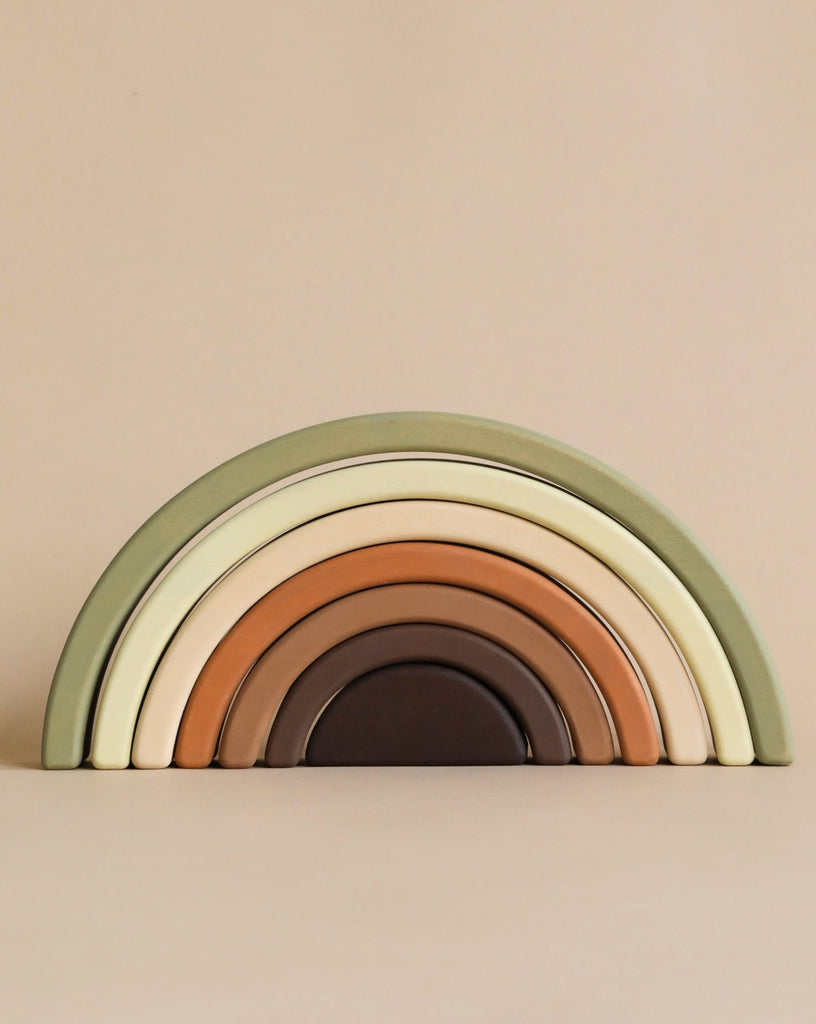 A Handmade Rainbow Stacker - Olive featuring seven arch segments in muted earth tones, ranging from light green, cream, tan, and brown. Made with non-toxic paint, the piece is set against a plain beige background.