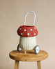 A whimsical Olli Ella Rattan Mushroom Luggy on wheels, featuring a red top with white polka dots and a white lower section, displayed on a wooden stool against a beige background.