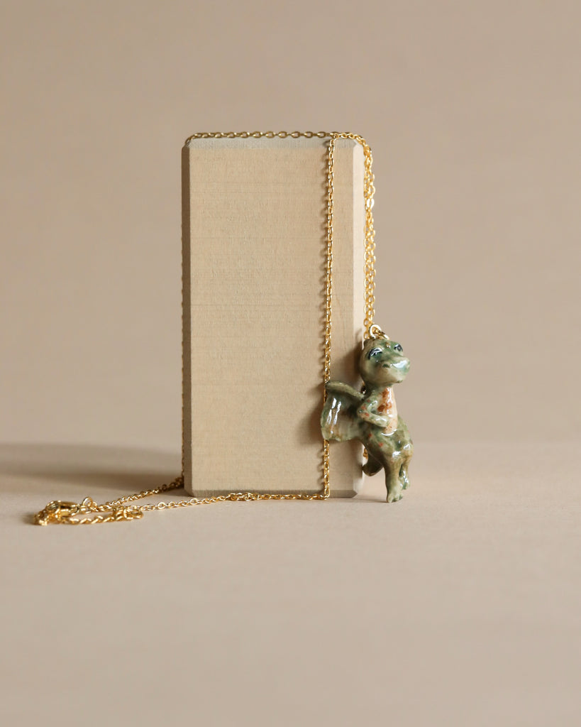 A handcrafted Baby Dragon Necklace attached to a gold chain, using it to lean against a standing beige book on a plain background.