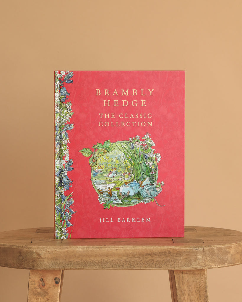 Brambly Hedge: The Classic Collection by Jill Barklem" is a red hardcover book with floral and nature illustrations on the cover, standing upright on a wooden stool against a beige background.