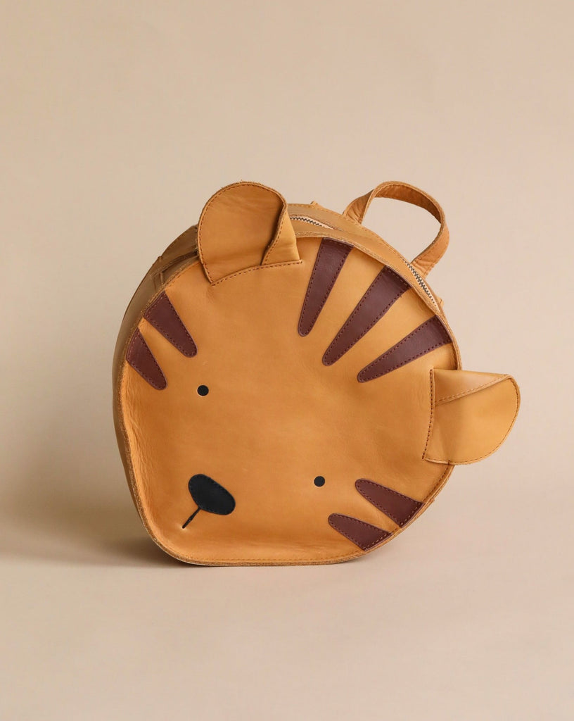 A children's backpack designed to look like a tiger's face, featuring ears and stripes in a Donsje School Leather Backpack - Tiger design, displayed against a neutral beige background.
