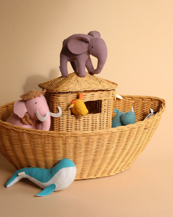 A beige Braided Ark filled with colorful plush animals, including a pink elephant, a blue bird, and others, with a large purple elephant sitting on top of the basket lid against a beige.